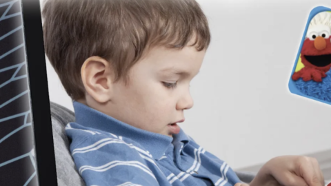 The image shows a young child, dressed in a blue striped shirt, focused on using a tablet or similar device. In the upper right corner of the image, there is a small icon featuring Elmo and Cookie Monster, characters from Sesame Street, suggesting that the child might be interacting with a Sesame Street app, possibly "Sesame Street Alphabet Kitchen." The background is plain, keeping the focus on the child and the device